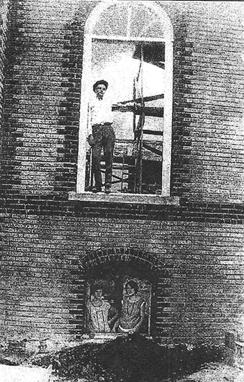 Brick church under construction with white man standing in upper story window frame and two white women looking out the bottom story window frame