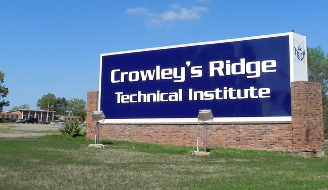 Large blue sign with white text and lights on grass with Brick building with parking lot behind it