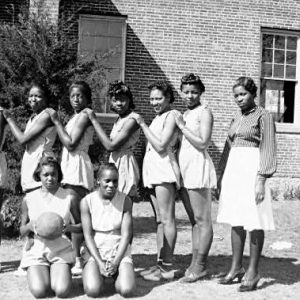 nine African American teen girls posing in basketball uniforms alongside an African American woman in a dress