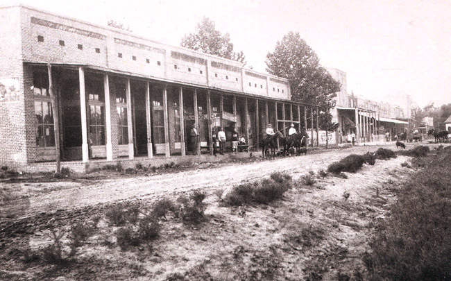 Group of white men standing and two white men on horseback at brick storefront with covered walkway on dirt road