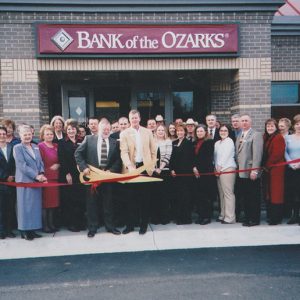 Men and women standing in front of bank with ribbon and one man holding large novelty scissors