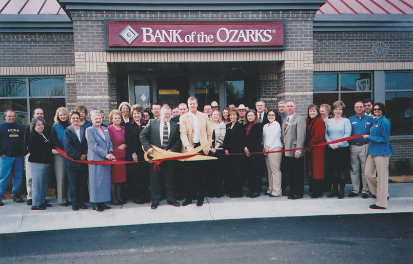 Men and women standing in front of bank with ribbon and one man holding large novelty scissors