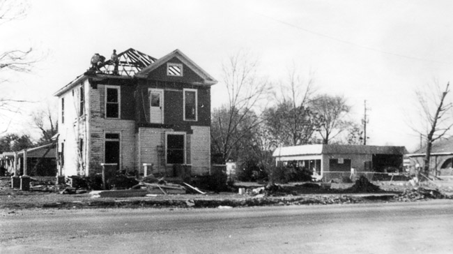 Two-story house with roof partially destroyed sitting on street with other debris visible and other structures in the distance