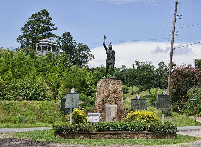 Statue of Native American man on stone pedestal in flower bed with historical markers surrounding it and gazebo on hill in the background