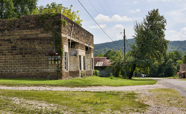 Side view of brick building and single-story buildings on rural road with tree-covered mountain in the background
