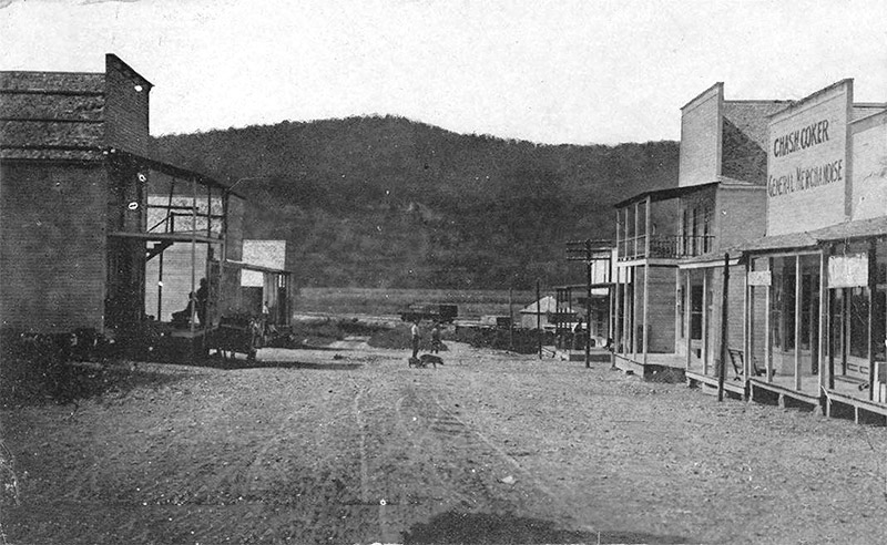 Storefront buildings with covered porches on dirt street with hill in the background