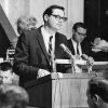 White man with glasses in suit speaking into microphones at lectern with white men and women sitting behind him
