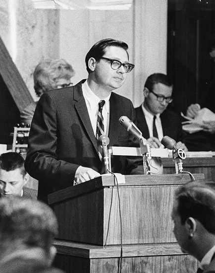 White man with glasses in suit speaking into microphones at lectern with white men and women sitting behind him