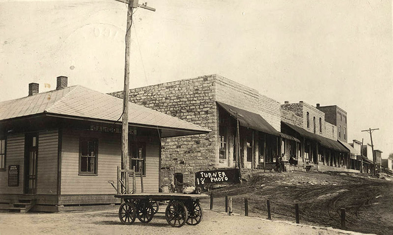 Train depot building with cart next to row of brick storefronts on dirt street
