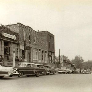 Cars parked outside brick storefronts on street with group of men sitting outside drug store