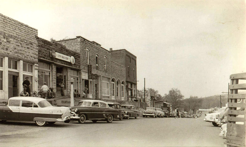 Cars parked outside brick storefronts on street with group of men sitting outside drug store