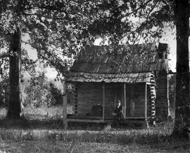 White man sitting on porch of log cabin between two trees