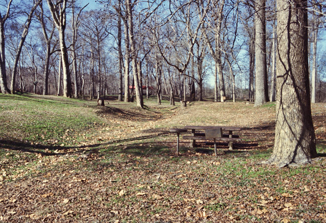 Concrete picnic table metal grill and round table in field with trees and red roof in the background