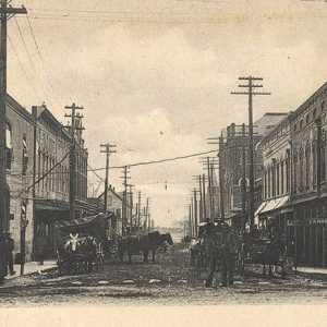 Horse drawn carriages on dirt street between rows of multistory buildings under power lines