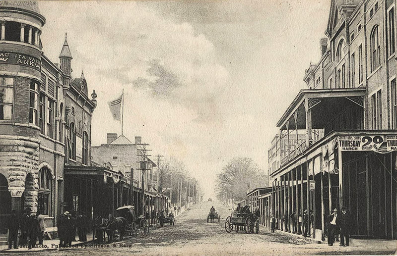Horse drawn carriages on dirt road with multistory buildings on both sides with covered entrances