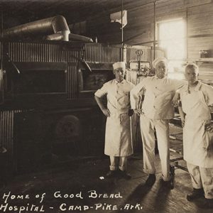 Four white men in matching uniforms standing in bakery with large oven and bread racks behind them
