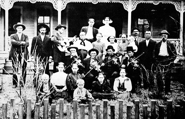 Group of white musicians with their instruments accompanied by white women and children inside fence with house behind them