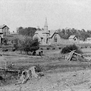 Bridge over creek in field with trees with multistory houses and church with steeple in the background