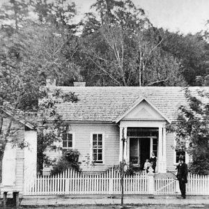 Single-story house with white woman and children standing on porch while white man and suit stands at fence