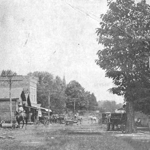 Men on horseback and horse drawn carriages on town street with storefronts on both sides