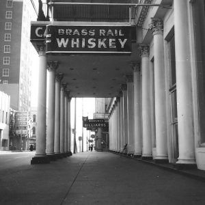 "Brass Rail Whiskey" neon sign on covered walkway supported by columns on sidewalk outside hotel building