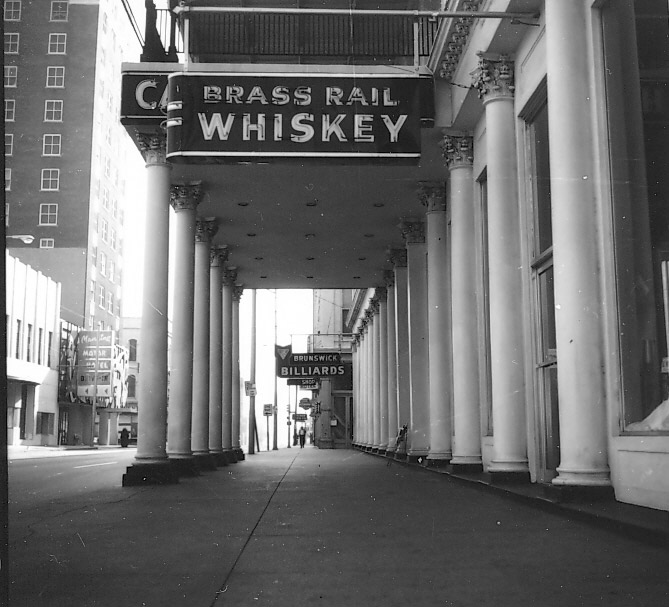 "Brass Rail Whiskey" neon sign on covered walkway supported by columns on sidewalk outside hotel building