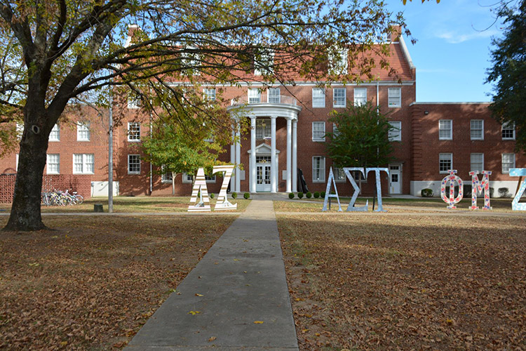 Multistory brick building with round covered porch with columns and Greek letters in front yard with trees