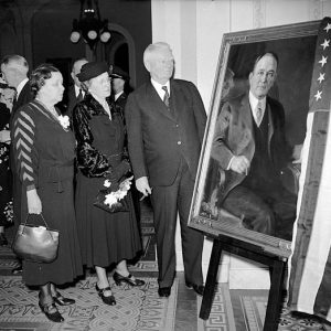 Old white man with two white women looking at a portrait or white man in suit sitting