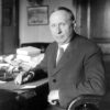 White man in suit and tie posing at his desk with pen in hand