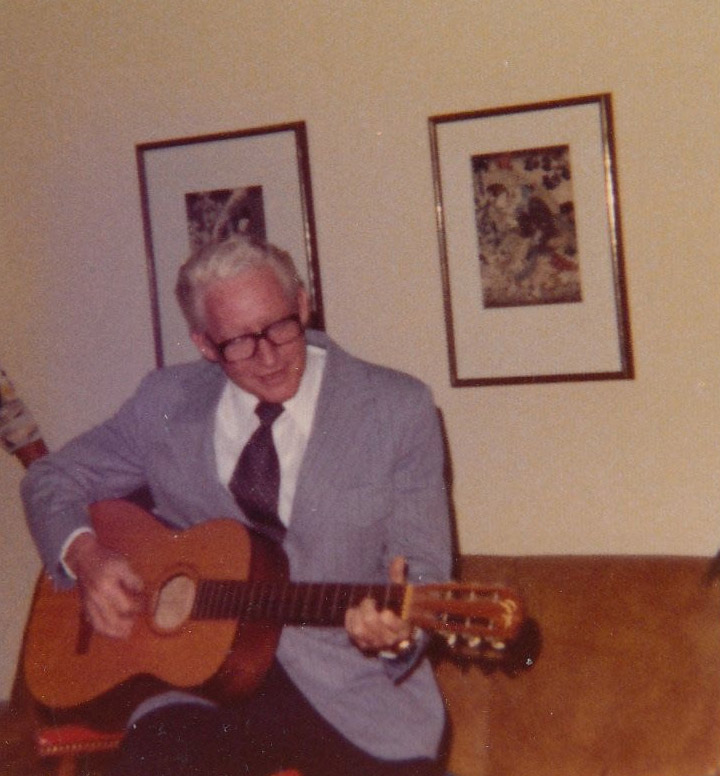 Old white man in suit and glasses playing an acoustic guitar on couch