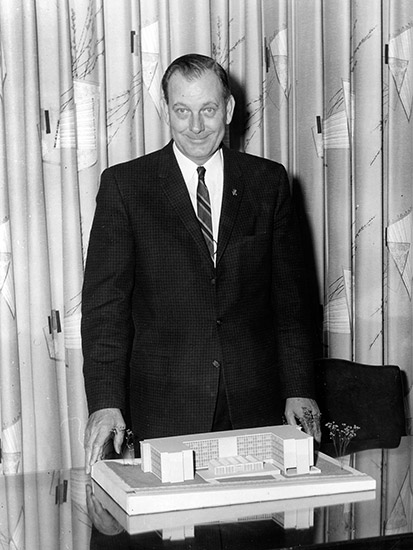 White man smiling in suit and tie with building model on table