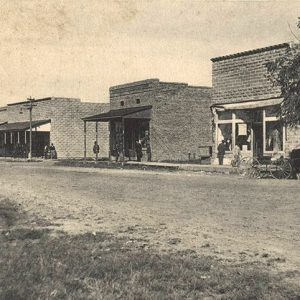 Single-story storefront buildings with covered porches on dirt street with horse drawn wagons and patrons