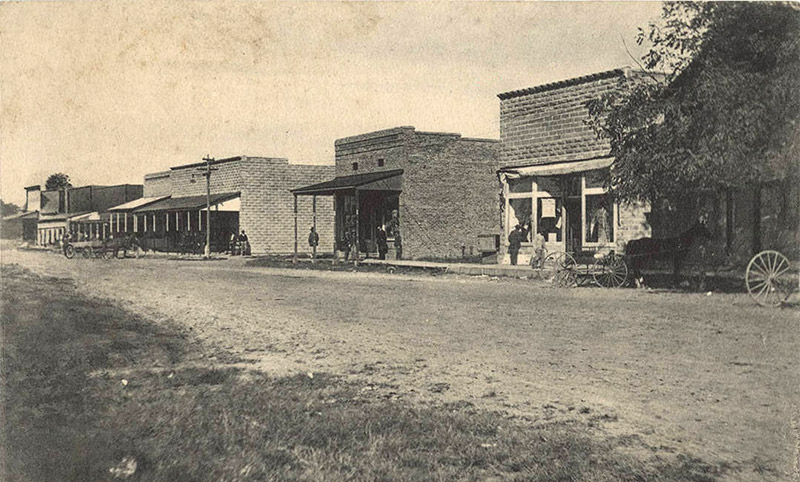 Single-story storefront buildings with covered porches on dirt street with horse drawn wagons and patrons