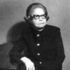 Old white woman in horned cat eye glasses and suit posing with arm on table