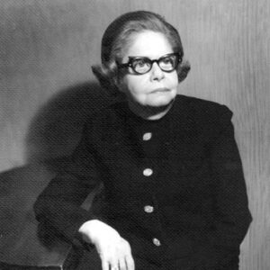 Old white woman in horned cat eye glasses and suit posing with arm on table