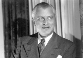 White man in suit and tie with curtains behind him
