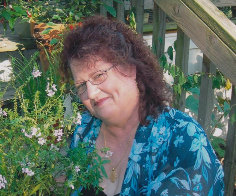 White woman with long hair and glasses in blue floral top surrounded by plants