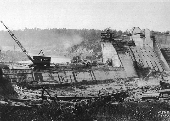 Concrete dam under construction with cranes and worker standing on bridge