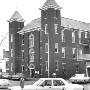 Three-story brick building with twin bell towers and street with parked cars