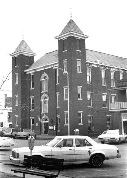 Three-story brick building with twin bell towers and street with parked cars