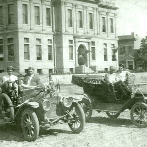 Four white men sitting in two old cars in front of large stone building