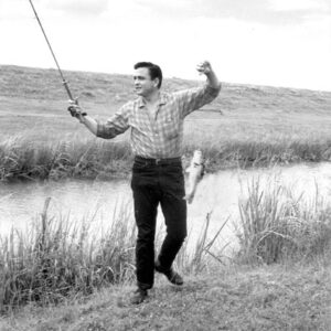 White man in shirt and jeans standing with fishing pole and fish on the line