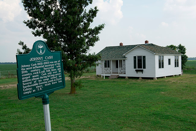 Single-story house with white paneling and historical marker sign on grass with tree