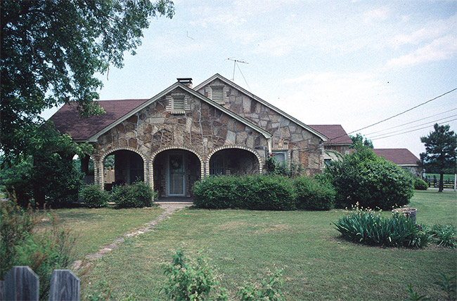 Stone house with arches in front with bushes tree and wooden fence and narrow stone walkway