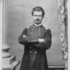 White man with mustache standing with arms crossed in military uniform