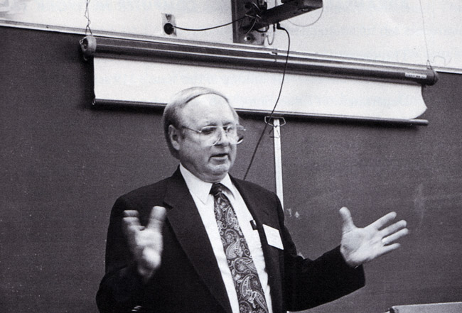 White man with glasses and suit speaking in chalkboard with pull-down screen