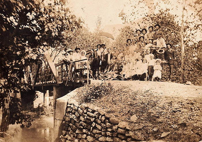 Crowd of white men women and children gathered on bridge over creek with concrete supports