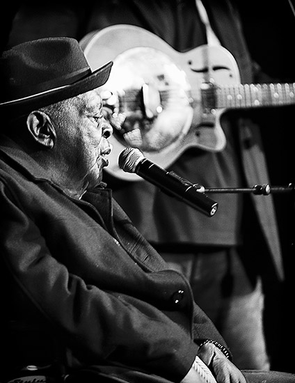 Seated African-American man singing into microphone on stage with guitarist in the background