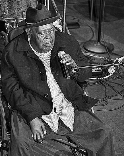 Older African-American man seated and singing into microphone on stage