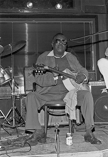 African-American man with sunglasses sitting with acoustic guitar on stage